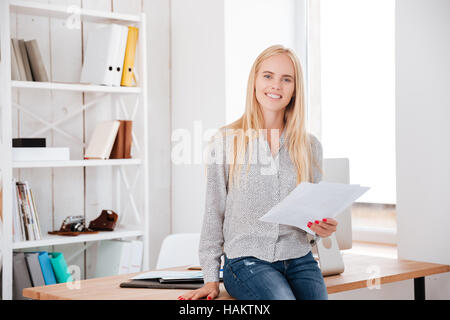 Lächelnde junge Geschäftsfrau hält Dokumente und sitzt auf dem Schreibtisch Stockfoto