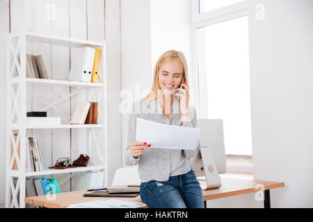 Glücklich lächelnde Frau, telefonieren mit Handy und halten Dokument beim Sitzen an ihrem Arbeitsplatz Stockfoto