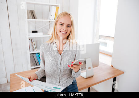 Cheeerful junge Geschäftsfrau mit Handy im stehen an ihrem Arbeitsplatz Stockfoto