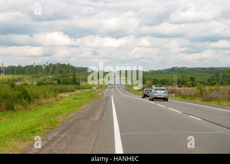 Asphaltierte Straße durch grüne Felder und die Autos drauf. Bewaldeten Bergen im Hintergrund. Sommertag mit weißen Wolken. Stockfoto
