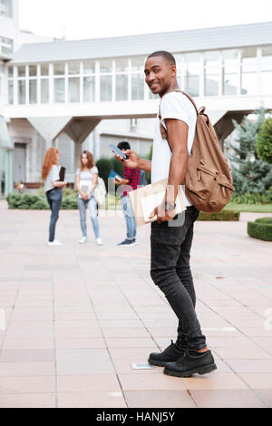 Lächelnd afroamerikanischen jungen Mann Student mit Rucksack zu Fuß auf dem campus Stockfoto