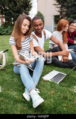 Multiethnische Gruppe glückliche junge Menschen Buch zu lesen und mit Laptop auf Rasen im freien Stockfoto