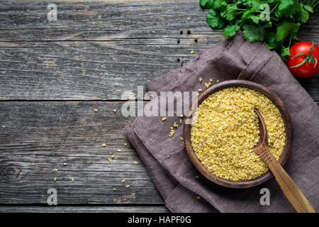 Bulgur (trockene Weizenkörner) in Holzschale, frische Petersilie, Tomaten und Gewürzen auf Holztisch Hintergrund. Draufsicht mit Textfreiraum Stockfoto