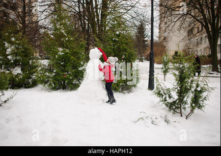 Das Mädchen im schulpflichtigen Alter baut einen Schneemann. Im Erdgeschoss gibt es eine Menge Schnee. Das Mädchen in eine rote Jacke und eine graue Strickmütze steht in der Nähe eines großen snowm Stockfoto