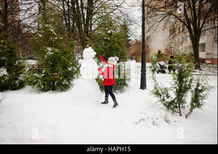 Das Mädchen im schulpflichtigen Alter baut einen Schneemann. Im Erdgeschoss gibt es eine Menge Schnee. Das Mädchen in eine rote Jacke und eine graue Strickmütze steht in der Nähe eines großen snowm Stockfoto