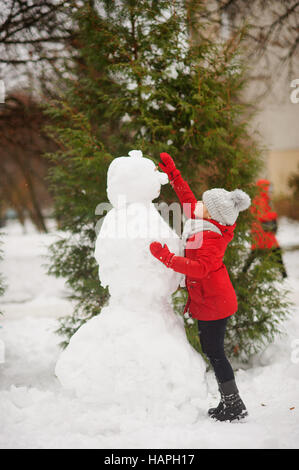 Das Mädchen im schulpflichtigen Alter baut einen Schneemann. Im Erdgeschoss gibt es eine Menge Schnee. Das Mädchen in eine rote Jacke und eine graue Strickmütze steht in der Nähe eines großen snowm Stockfoto