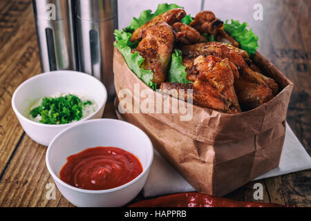 leckere gegrillte Hähnchenflügel mit Knoblauch und Tomaten-Sauce mit Salat in Essen Papiertüte auf hölzernen rustikalen Hintergrund Draufsicht, Witi Exemplar Stockfoto