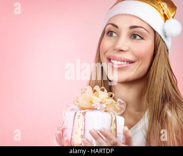 Porträt von einem netten Santa Mädchen mit goldenen festliche Hut mit Geschenkbox in Händen über rosa Hintergrund, schöne Lächeln Santa Helfer, Textfreiraum Stockfoto