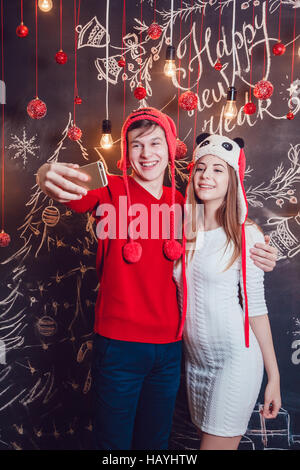 Glücklich Couplein lustige Hüte stehend umarmt und machen Selfie auf einem dunklen Hintergrund mit Weihnachten Muster. Neues Jahr. Stockfoto