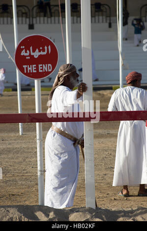 Mann mit weißem Bart steht hält ein Pole neben ein Zeichen auf der Ziellinie von einem Kamelrennen in Oman Stockfoto