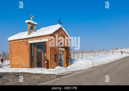 Kleine Kapelle am Straßenrand und Weinbergen bedeckt mit Schnee unter blauem Himmel im Piemont, Norditalien. Stockfoto