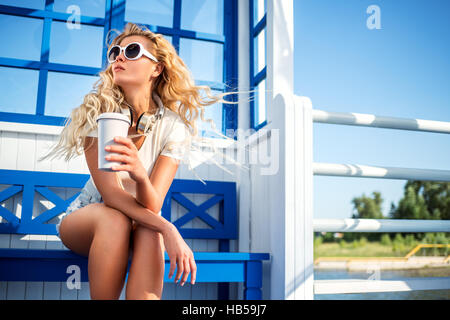 Schöne junge Frau mit Kopfhörern Musik und Take away Kaffeetasse, auf der Bank sitzen und warten auf ein Wassertaxi. Stockfoto