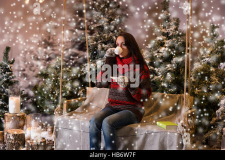 Frau sitzt auf einer Schaukel mit einer Decke unter den Taschenlampen und Drinkig eine Tasse Kaffee in einem verschneiten Park mit Fichten, tragen rote Wolle Stockfoto