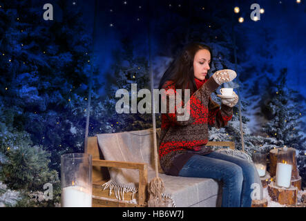 Frau sitzt auf einer Schaukel mit einer Decke unter den Taschenlampen und hält eine Tasse Kaffee in einem verschneiten Park am Abend tragen rote Wolle sw Stockfoto