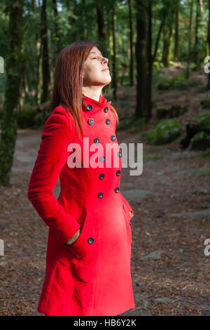 Frau, genießen die Wärme des Sonnenlichts auf einen Wald tragen einen roten Mantel winter Stockfoto