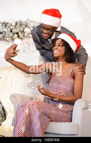 Das junge ziemlich afrikanische Mädchen im Christmas Cap im Sessel sitzen und Selfies mit ihren hübschen Freund machen Stockfoto