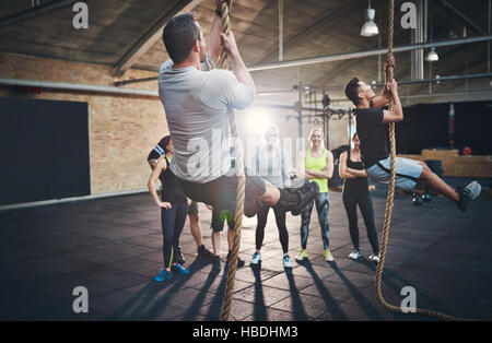 Zwei Männer in kurzen Hosen Dicke Klettertau während Fitness-Übungen in Cross-Fit Training mit anderen Erwachsenen beobachtete sie von hinten Stockfoto