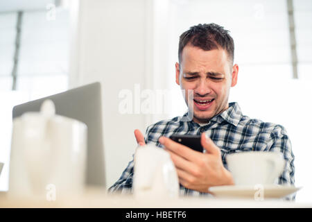 Schlechte Nachrichten. Zorniger junger Mann hält ein Mobiltelefon und Grimassieren im café Stockfoto