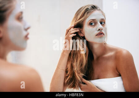 Weibchen mit kosmetische Gesichtsmaske im Badezimmer. Frau in Blick in den Spiegel mit Maske auf ihr Gesicht Handtuch gewickelt. Stockfoto