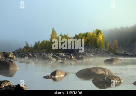 Steinen im Wasser im Vordergrund und eine kleine Insel im Nebel und wenig Sonne im Hintergrund, Bild aus Nordschweden. Stockfoto