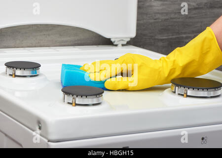 Zarte Frauenhand in gelb Kautschuk Schutzhandschuh Reinigung weißes Gas Herd mit blauen Schwamm auf grauem Hintergrund Stockfoto