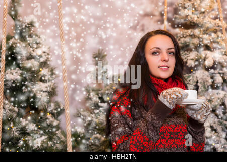 Porträt der lächelnde junge Frau unter den Taschenlampen sitzt und hält eine Tasse Kaffee in einem verschneiten Park mit Fichten, tragen rote Wolle Stockfoto