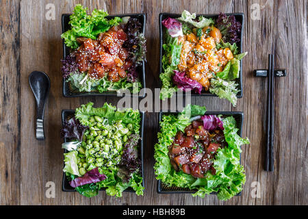 Draufsicht auf 4 Poke-Schüsseln mit Essstäbchen und einem Löffel auf einer Holzfläche. 3 Fischoptionen und eine vegetarische Option. Stockfoto