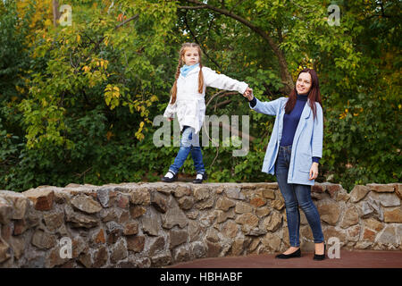 Mutter und Tochter im Herbst Park. Mama hilft ihrer Tochter auf dem Stein Zaun gehen. Zeit mit der Familie. Stockfoto