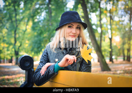 Wunderschöne stilvolle junge Frau tragen modische Kleidung. Porträt von Mädchen in schwarzen Hut sitzt auf der Bank im Park. Stockfoto