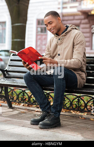 Foto des jungen afrikanischen glücklicher Mann ein Buch zu lesen, während Sie sitzen auf einer Bank an der Straße Stockfoto
