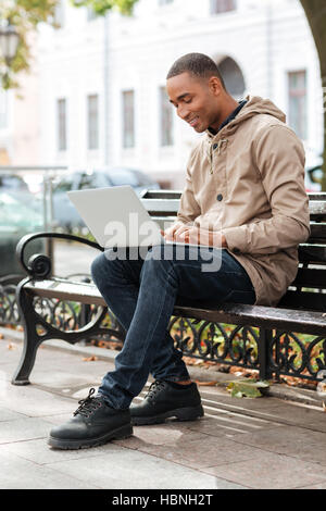 Foto von jungen afrikanischen Mann mit Laptop sitzt auf einer Holzbank und eingeben. Stockfoto