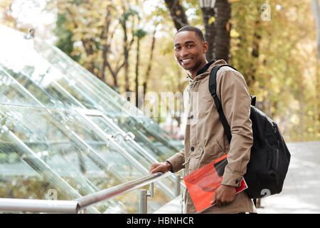 Foto von jungen afrikanischen fröhlichen Mann mit Rucksack stehen auf der Straße halten Sie Buch und schauen in die Kamera. Stockfoto