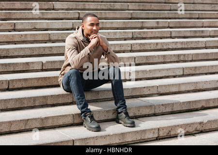Bild der jungen afrikanischen fröhlicher Mensch sitzt auf der Treppe. Auf der Suche zur Seite. Stockfoto