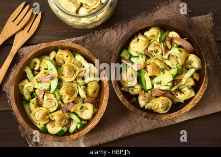 Gebackener Käse Tortellini oder Bauchnabel Pasta mit Zucchini, Speck und Thymian in Holzschalen, overhead auf dunklem Holz mit natürlichem Licht fotografiert Stockfoto