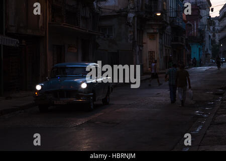 Scheinwerfer von einem klassischen Auto durchdringen die Dunkelheit auf einer Straße in Havanna Centro eines Morgens kurz nach Sonnenaufgang. Stockfoto