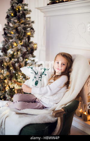 Mädchen zu Weihnachten, Weihnachtsbaum im Hintergrund, schöne junge Kind eingewickelt in eine Decke, sie lächelt Stockfoto