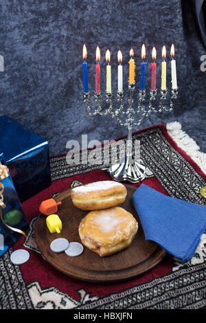 Hanukkah Menorah mit brennenden Kerzen, Geschenke, Dreidel und Gelee gefüllt Gebäck serviert auf einer Holzplatte für den Urlaub Stockfoto