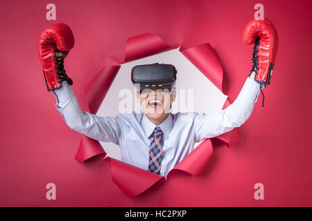 Porträt von lächelnden Jungunternehmer in virtual-Reality-Kopfhörer und Boxhandschuhe seine Hände in Loch gerissen auf rotem Papier Stockfoto