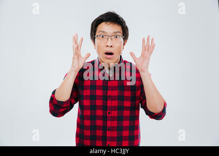 Foto von schockiert jungen asiatischen Mann gekleidet in lässigen Shirt in einem Käfig und tragen Brillen mit Händen gestikulieren. Isoliert auf weißem Hintergrund. Suchen Sie eine Stockfoto