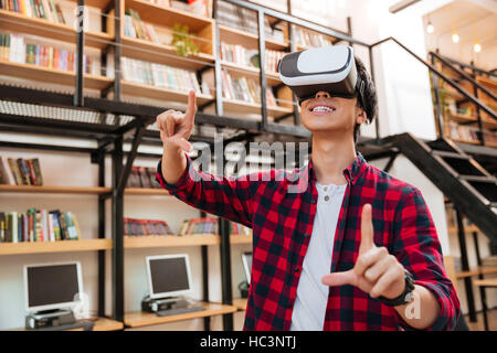 Glücklich Jüngling gekleidet im lässigen Hemd in einem Käfig und tragen virtual-Reality-Gerät in der Bibliothek. Stockfoto