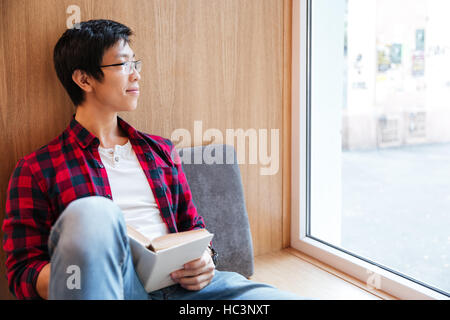 Bild des lächelnden asiatische Studentin Lesen eines Buches in Universitätsbibliothek auf Sofa sitzen. Suchen Sie im Fenster. Stockfoto