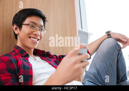 Bild des glücklichen asiatische Studentin im Chat in Universitätsbibliothek auf Sofa sitzen. Mit Blick auf Telefon. Stockfoto