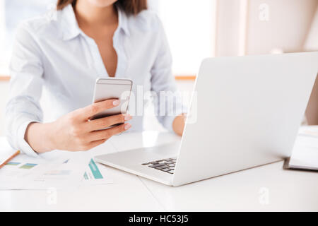 Bild der junge Geschäftsfrau gekleidet in weißen Hemd in ihrem Büro sitzt und hält Handy zugeschnitten. Stockfoto