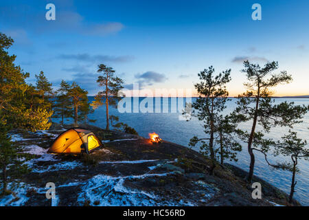 Lagerfeuer und Zelt in der Wildnis am See bei Sonnenuntergang, Schnee auf dem Boden Stockfoto