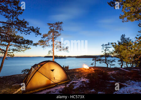 Lagerfeuer und Zelt in der Wildnis am See bei Sonnenuntergang, Schnee auf dem Boden Stockfoto