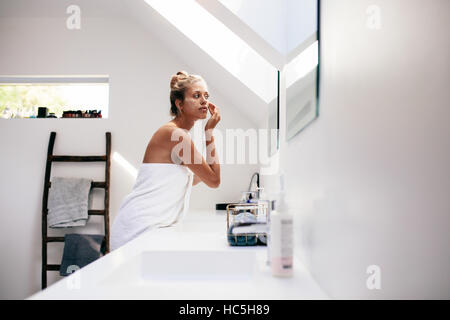 Junge Frau in Blick in den Spiegel und Auftragen der Creme im Gesicht Handtuch gewickelt. Frauen die Pflege ihrer Gesichtshaut im Badezimmer nach Bad. Stockfoto