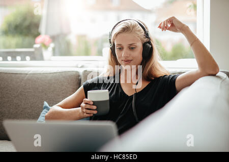 Schöne junge Frau mit Kopfhörern hält eine Tasse Kaffee und Blick auf Laptop. Frau sitzt auf der Couch im Wohnzimmer zu Hause.