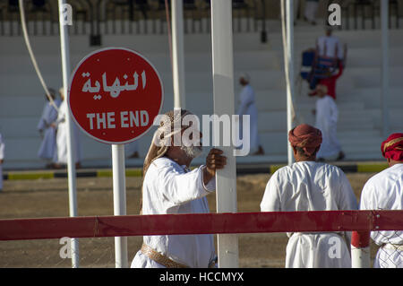 Mann mit weißem Bart steht hält ein Pole neben ein Zeichen auf der Ziellinie von einem Kamelrennen in Oman Stockfoto