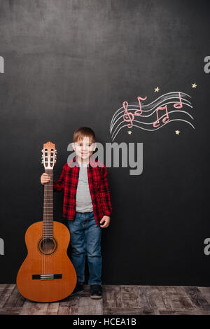 Foto von niedlichen kleinen Jungen halten die Gitarre im stehen in der Nähe von schwarzen Tafel mit musikalisch Zeichnungen. Stockfoto