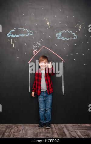 Glückliches kleines Kind stehen im Haus an der Tafel mit Zeichnungen von einem Regen. Blick in die Kamera, während der Daumen hoch Geste zu machen. Blick in die Kamera. Stockfoto